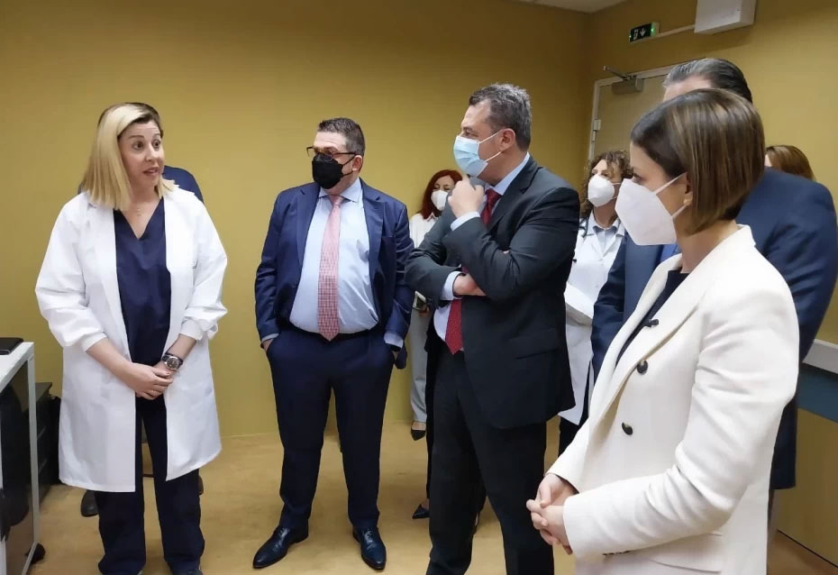 Επίσκεψη επισήμων σε νοσοκομειακό χώρο: γιατρός με λευκή ποδιά συζητά με ομάδα ατόμων με κουστούμια, οι περισσότεροι φορούν μάσκα προσώπου